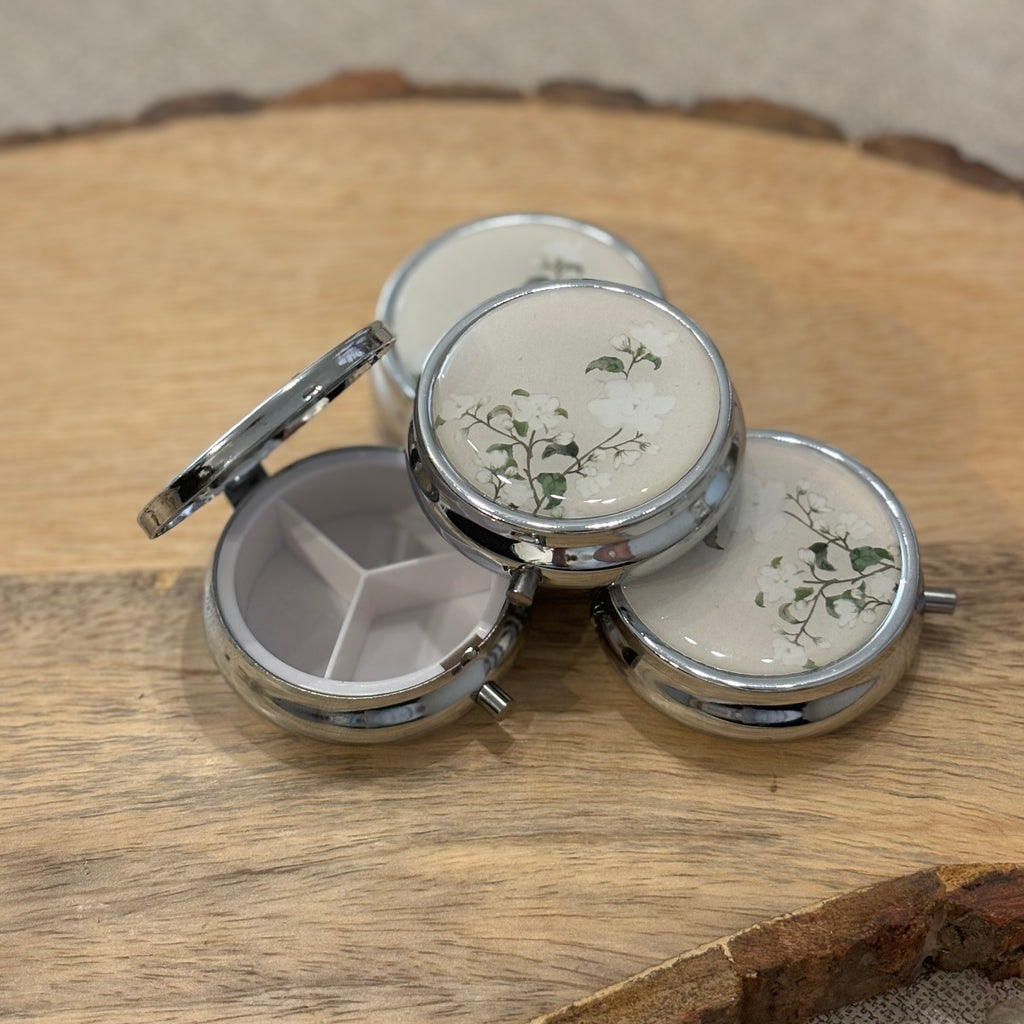 Multiple pill boxes with white flower design. One is open to show the inside compartment which is split into thirds. 