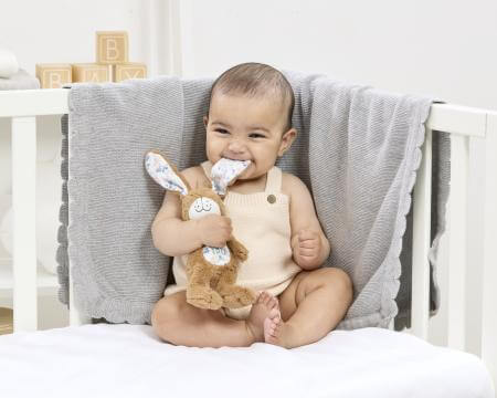 Baby sitting in a crib holding a plush toy, wearing a onesie with bunny ears.