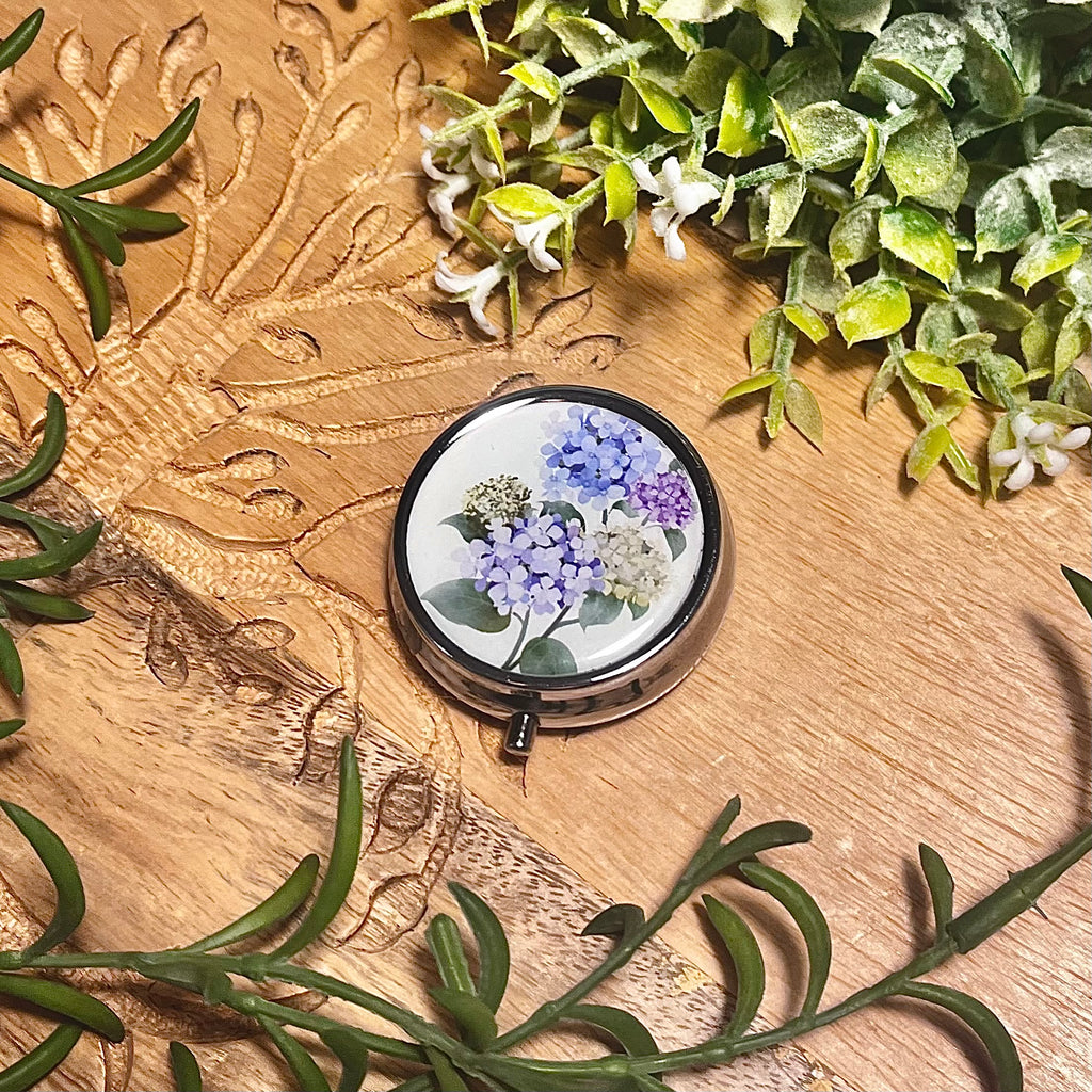Round silver pill box with cream top and blue, purple, and green flowers on. 