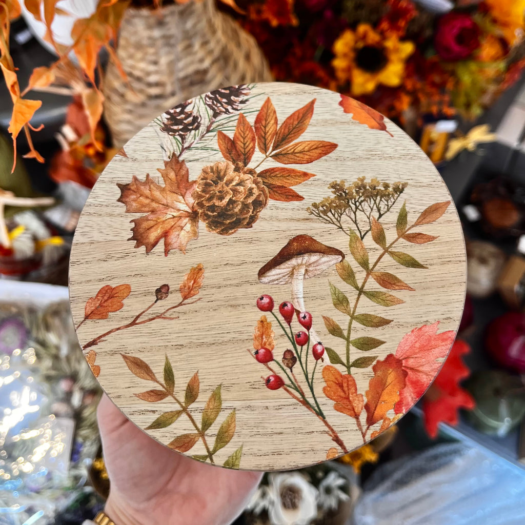 Round wooden candle lid with autumn-themed design held in a hand against a blurred background.