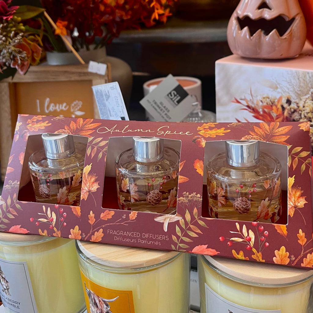 Set of three fragranced diffusers in a decorative box with autumn-themed design, surrounded by candles and pumpkins.