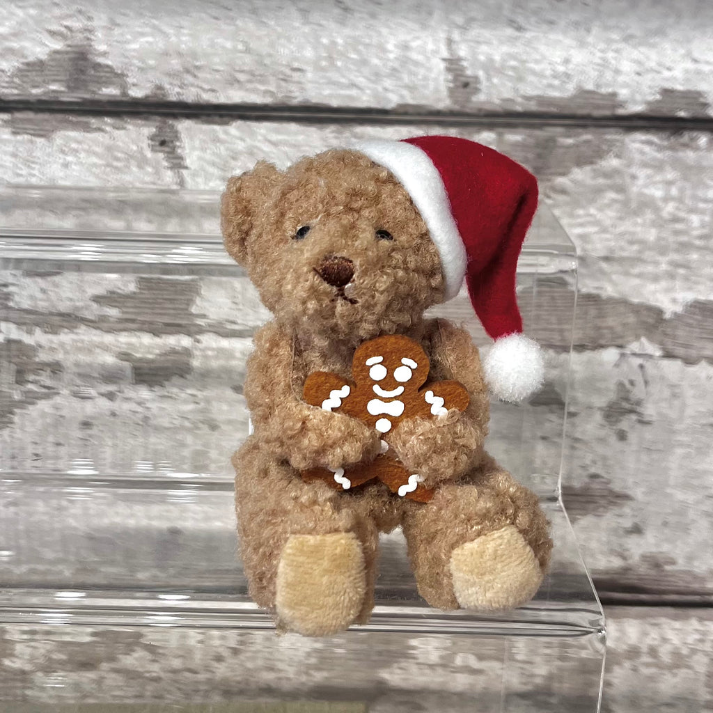 Teddy bear wearing a Santa hat holding a gingerbread man toy on a clear stand with a wooden background.