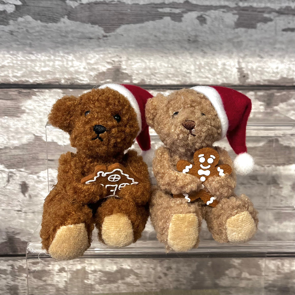 Two teddy bears wearing Santa hats, one with a gingerbread man and the other with a gingerbread house, on a wooden surface.