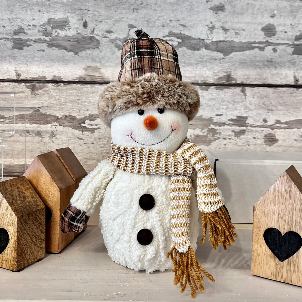 Decorative snowman with a plaid hat and knitted scarf in front of wooden houses with heart designs.