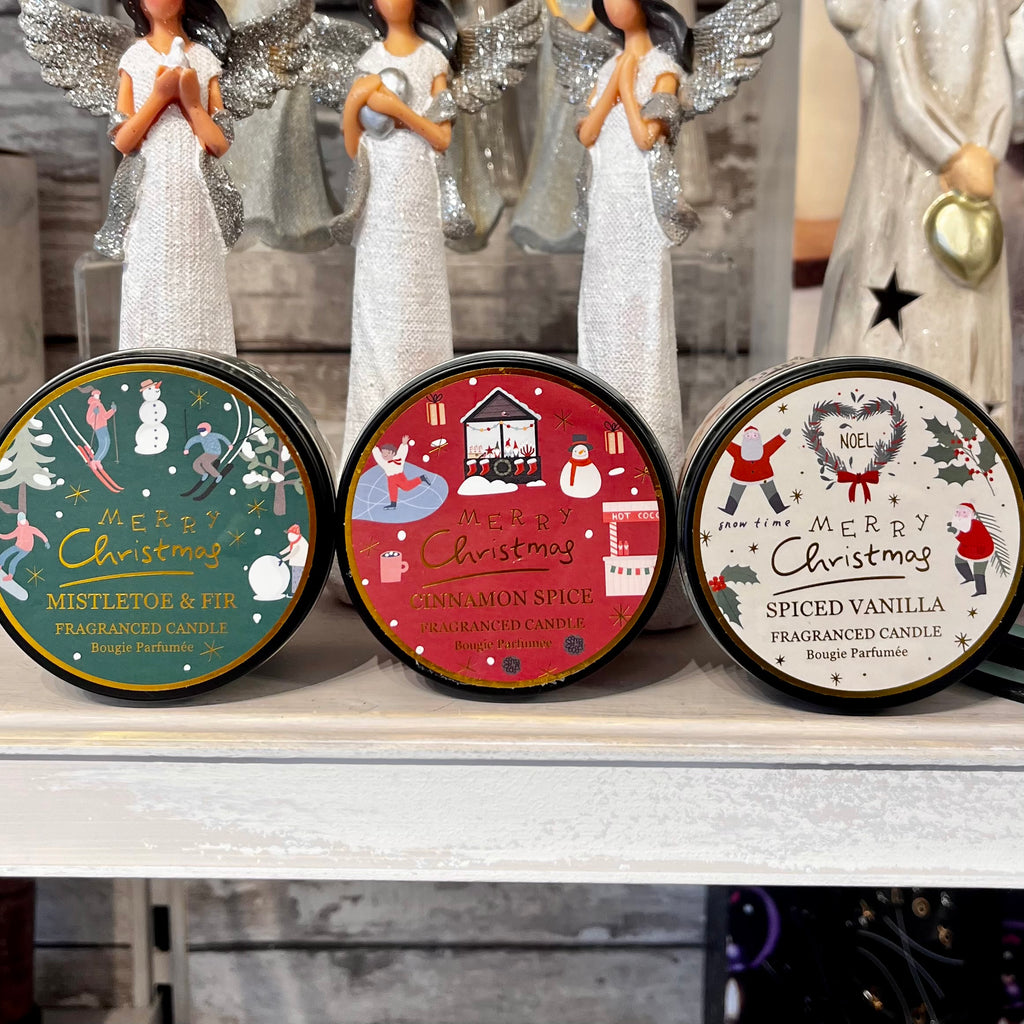 Three Christmas-themed candles with decorative labels on a shelf, accompanied by angel figurines.