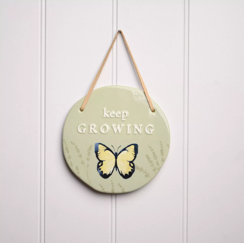 Green circle hanging plaque with darker green monochrome reeds on the bottom half of the plaque with a blue and yellow butterfly over the top of them. Above the butterfly is the quote 'keep growing' in a serif font