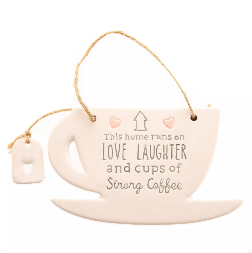 White ceramic hanging decoration in the shape of a mug on a coaster with ceramic teabag charm with heart cutout attached by twine. Features the quote 'This home runs of love, laughter, and cups of strong coffee'