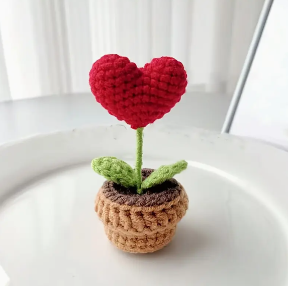 knitted plant with green stem. two leaves and a red heart in place of a flower. Sits on top of a knitted brown plant pot.