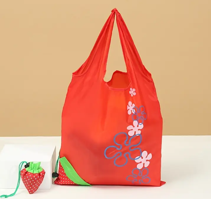 Red shoulder bag with cartoon flower shapes in a line leading up to the top. On the corner is the strawberry design with a drawstring attachment where the bag gets folded into .